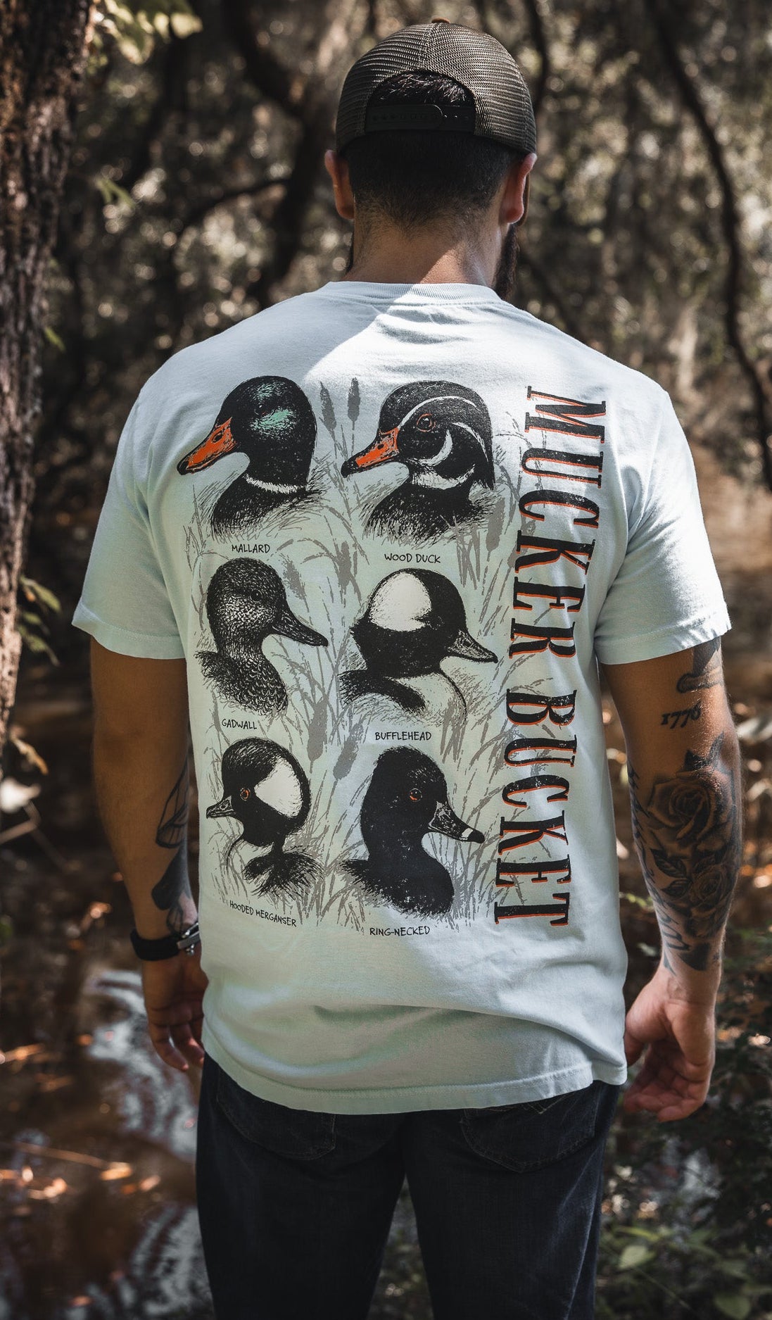 A man in a cap stands by a wooded stream, wearing the Duck DNA Premium Tee by Mucker Bucket—a relaxed fit light blue shirt featuring illustrations and names of five duck species with “Mucker Bucket” printed vertically on the back.