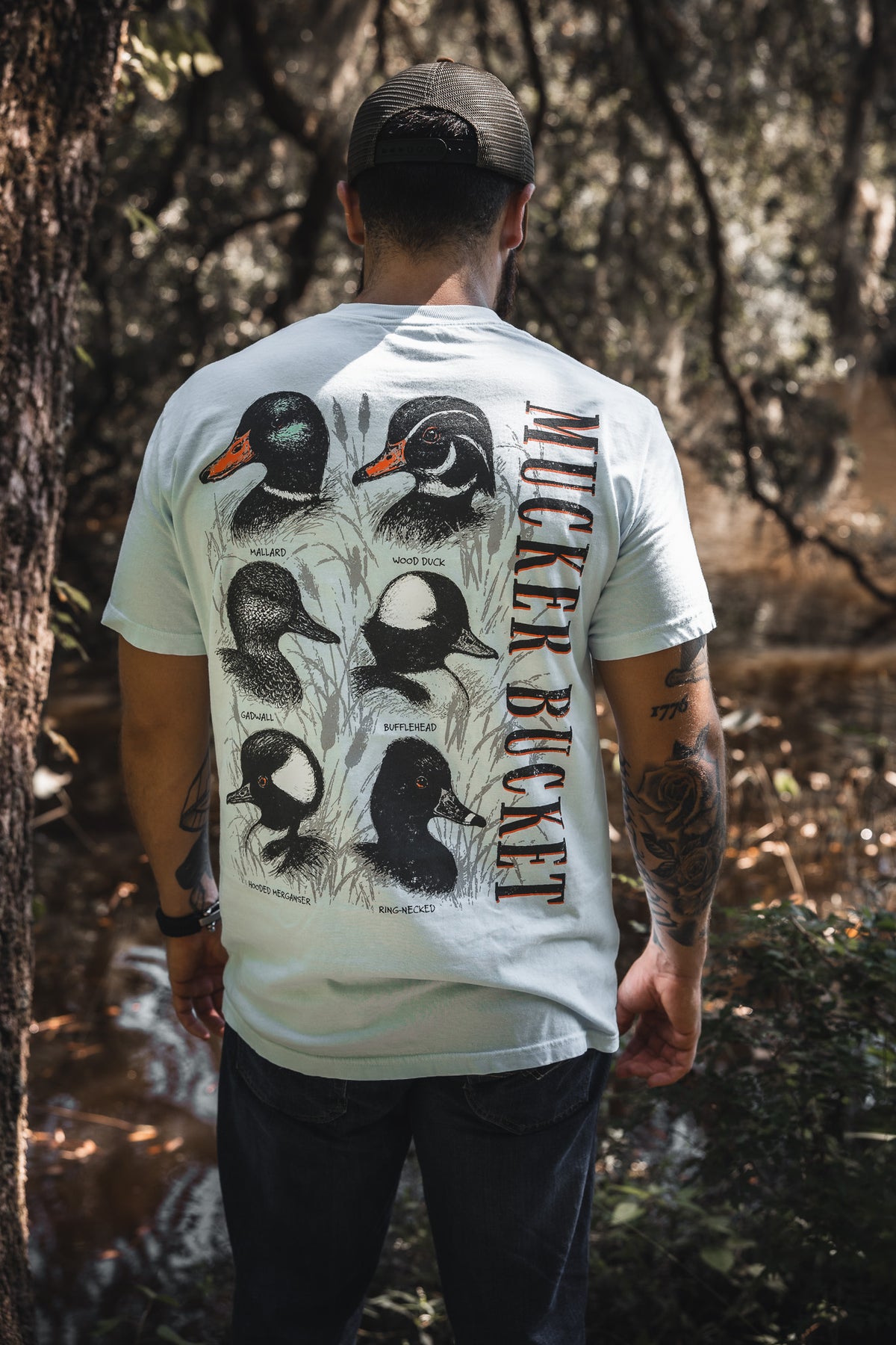 A man in a cap stands by a wooded stream, wearing the Duck DNA Premium Tee by Mucker Bucket—a relaxed fit light blue shirt featuring illustrations and names of five duck species with “Mucker Bucket” printed vertically on the back.