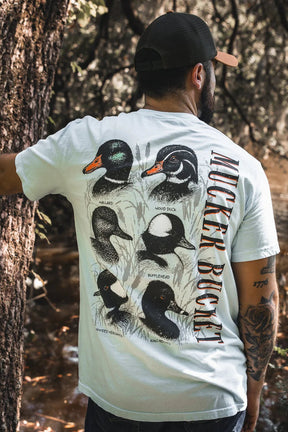 A man stands by a tree, facing away, wearing the Duck DNA Premium Tee from Mucker Bucket. The relaxed fit white shirt features illustrated duck species and "MUCKER BUCKET" vertically on the back, with a stream and trees in the background.