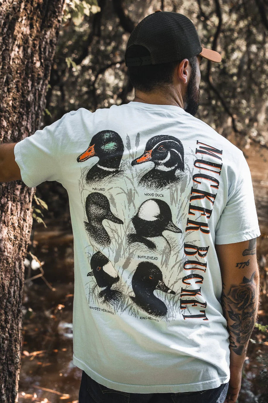 A man stands by a tree, facing away, wearing the Duck DNA Premium Tee from Mucker Bucket. The relaxed fit white shirt features illustrated duck species and "MUCKER BUCKET" vertically on the back, with a stream and trees in the background.