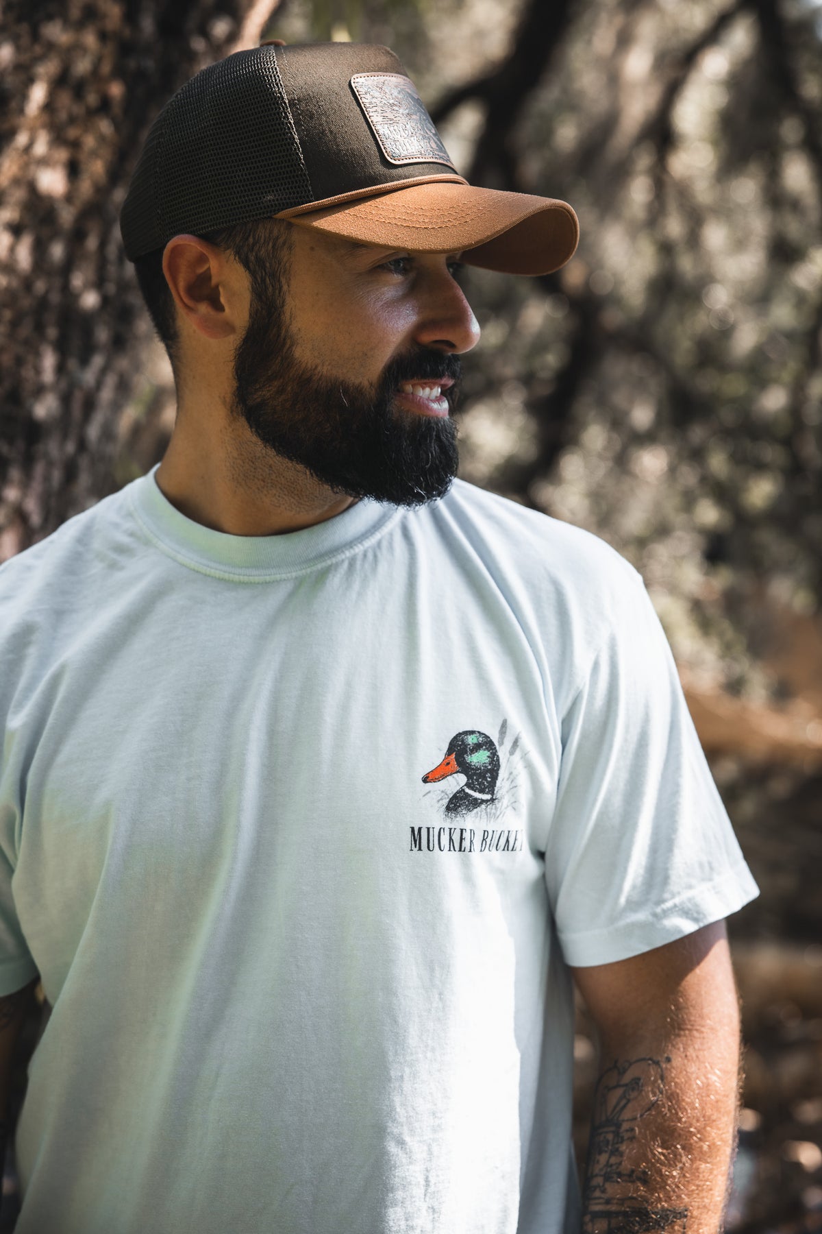 A bearded man in a brown cap smiles outdoors, wearing the Mucker Bucket Duck DNA Premium Tee—a light garment-dyed cotton shirt featuring a duck graphic and the words "Mucker Bucket.