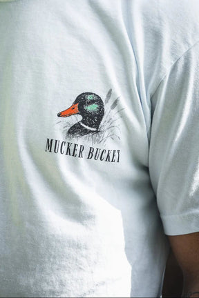 Close-up of the Duck DNA Premium Tee by Mucker Bucket, a garment-dyed light blue relaxed fit shirt with a duck head illustration and "MUCKER BUCKET" text. Part of the wearer's arm is partially visible.