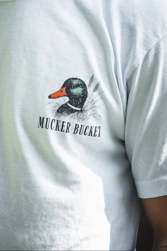 Close-up of the Duck DNA Premium Tee by Mucker Bucket, a garment-dyed light blue relaxed fit shirt with a duck head illustration and "MUCKER BUCKET" text. Part of the wearer's arm is partially visible.