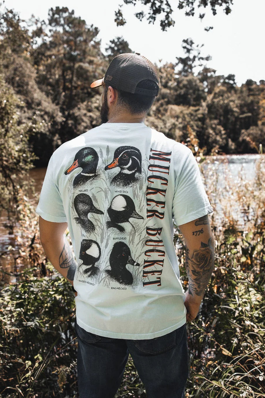 A man stands by a river wearing the Duck DNA Premium Tee from Mucker Bucket, featuring duck illustrations and “MUCKER BUCKET” on the back, paired with his tattoos, baseball cap, and a scenic backdrop of trees and water.