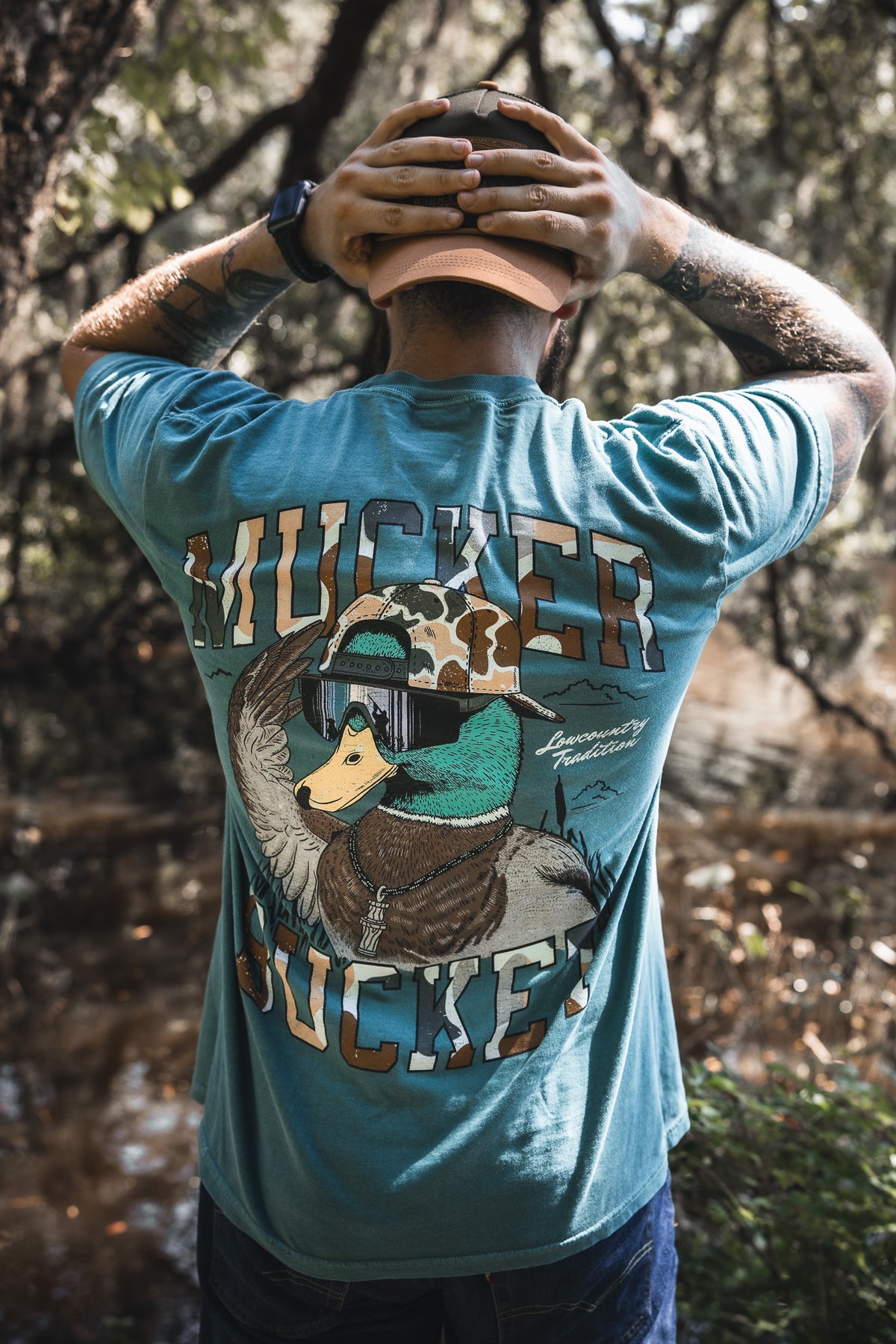 A tattooed person stands outdoors by trees and water, wearing the Mucker Bucket Cool Duck Premium Tee in blue, featuring a cartoon duck with helmet and goggles and "MUCKER SUCKER" text on the back.