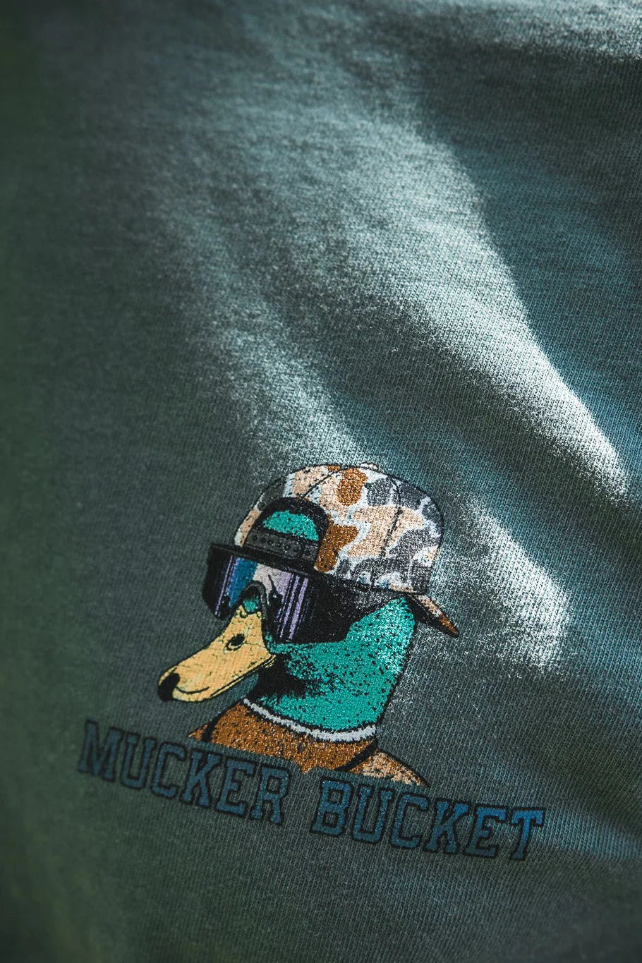 Close-up of the Mucker Bucket Cool Duck Premium Tee, featuring an illustrated duck in sunglasses and a camo hat, with "MUCKER BUCKET" printed below.