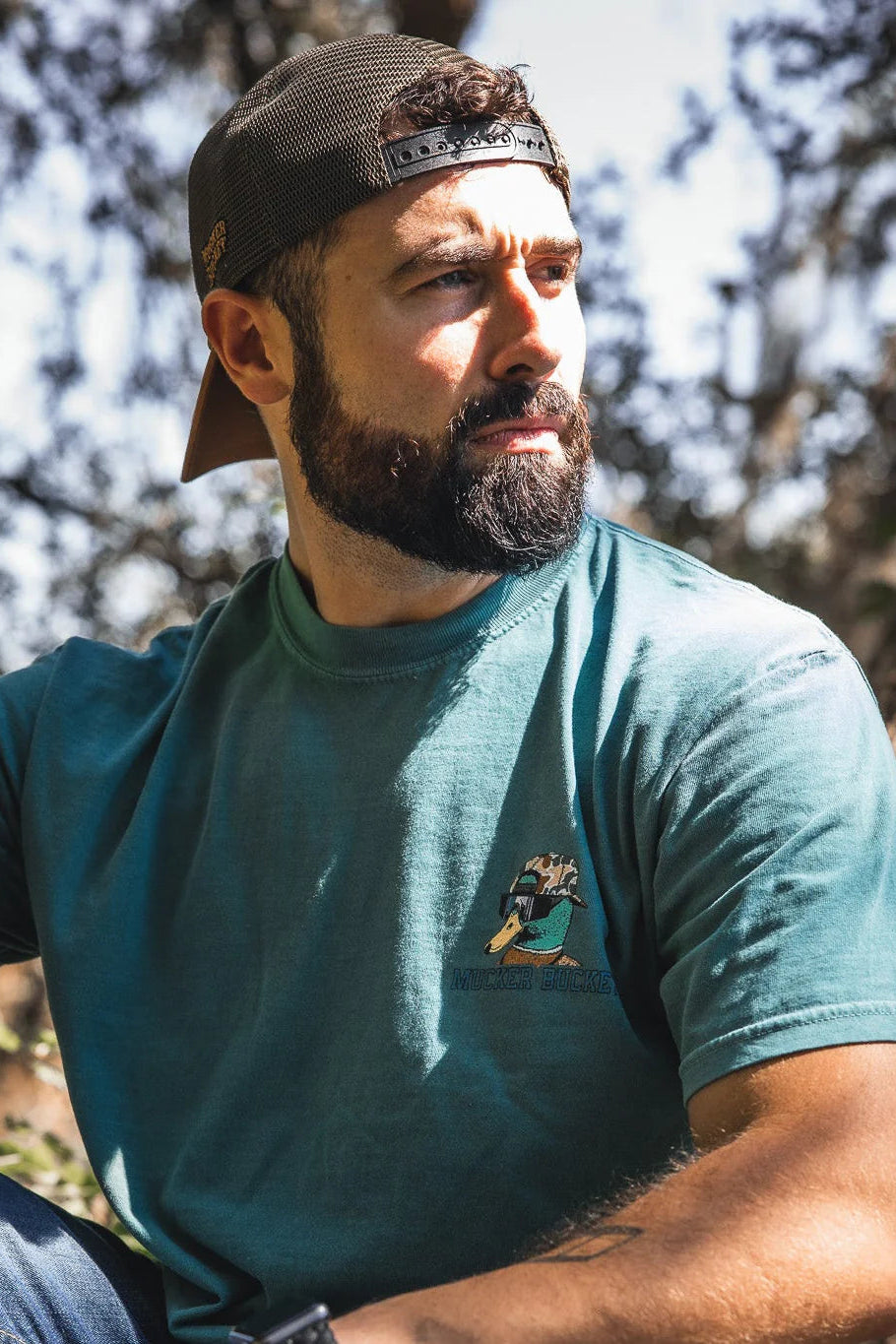A bearded man in a backward cap wears the Mucker Bucket Cool Duck Premium Tee—a green, garment dyed, relaxed fit shirt—while sitting outdoors in sunlight with trees and blurred foliage in the background.