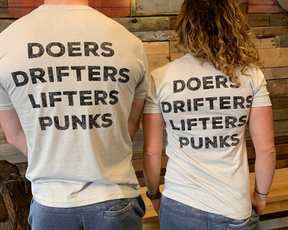 Two people, backs to the camera, wear the Doers Drifters Lifters Punks Unisex Tee by Tango Charlie Apparel. The bold slogan stands out against light fabric as they rep their crew in front of a rustic wooden wall.