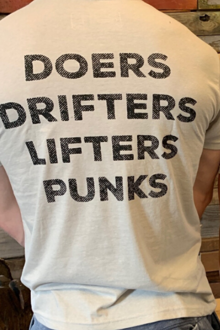 Two people, backs to the camera, wear the Doers Drifters Lifters Punks Unisex Tee by Tango Charlie Apparel. The bold slogan stands out against light fabric as they rep their crew in front of a rustic wooden wall.