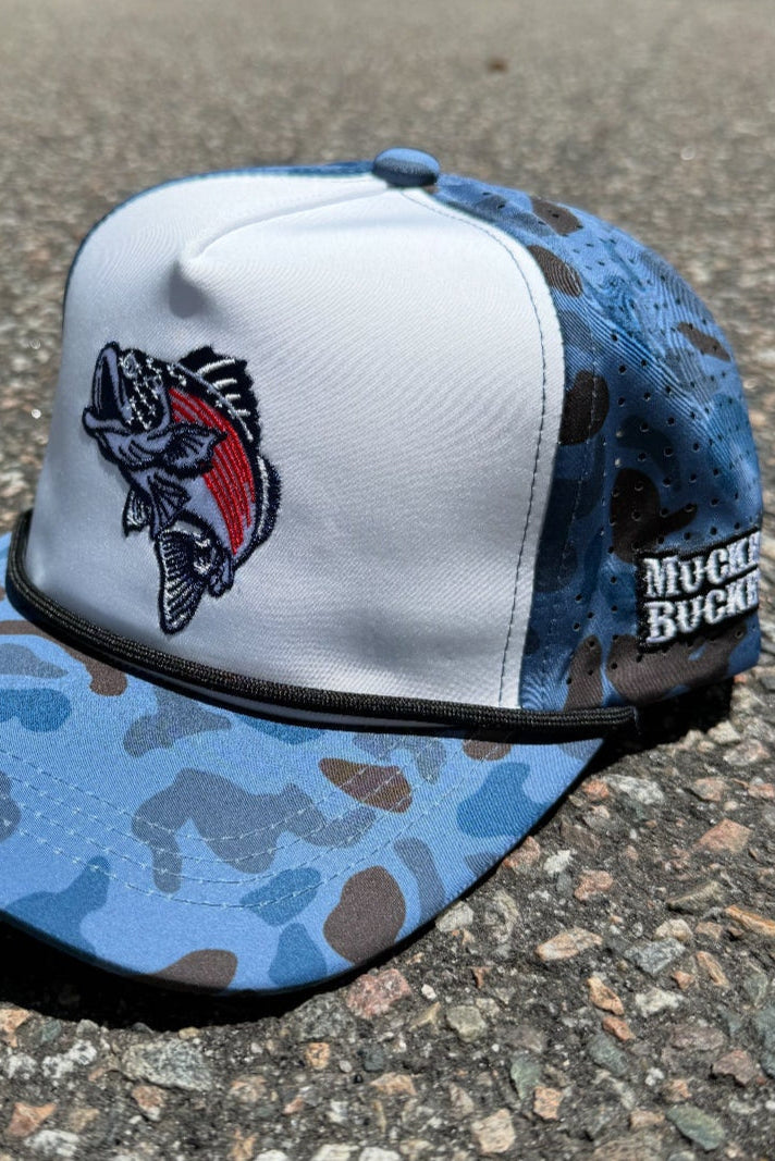 The Mucker Bucket Monster Bass 'Merica Water Camo Bucket, a blue and white camo snapback with an embroidered fish patch and "Muck Bucket" on the side, sits on rough rocks outdoors.