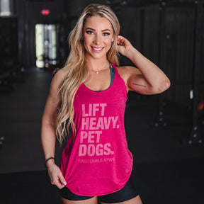 A smiling woman with long blonde hair stands in a dark, blurred gym, wearing the Lift Heavy. Pet Dogs.- Womens Pink Tank by Tango Charlie Apparel and black shorts. Her left hand touches her hair.
