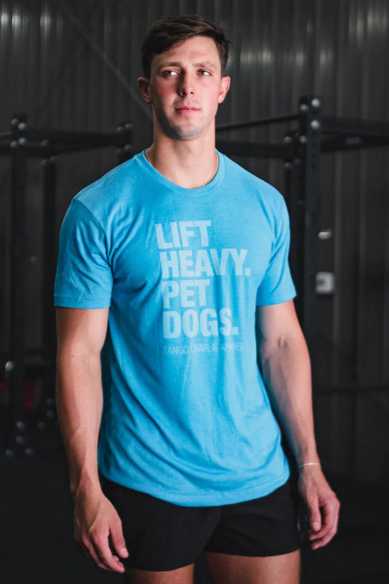 In a gym, a man wearing Tango Charlie Apparels turquoise Lift Heavy. Pet Dogs T-shirt with vintage-style ink and enjoying its ComfyAF tri-blend feel pairs it with black shorts amidst gym equipment.