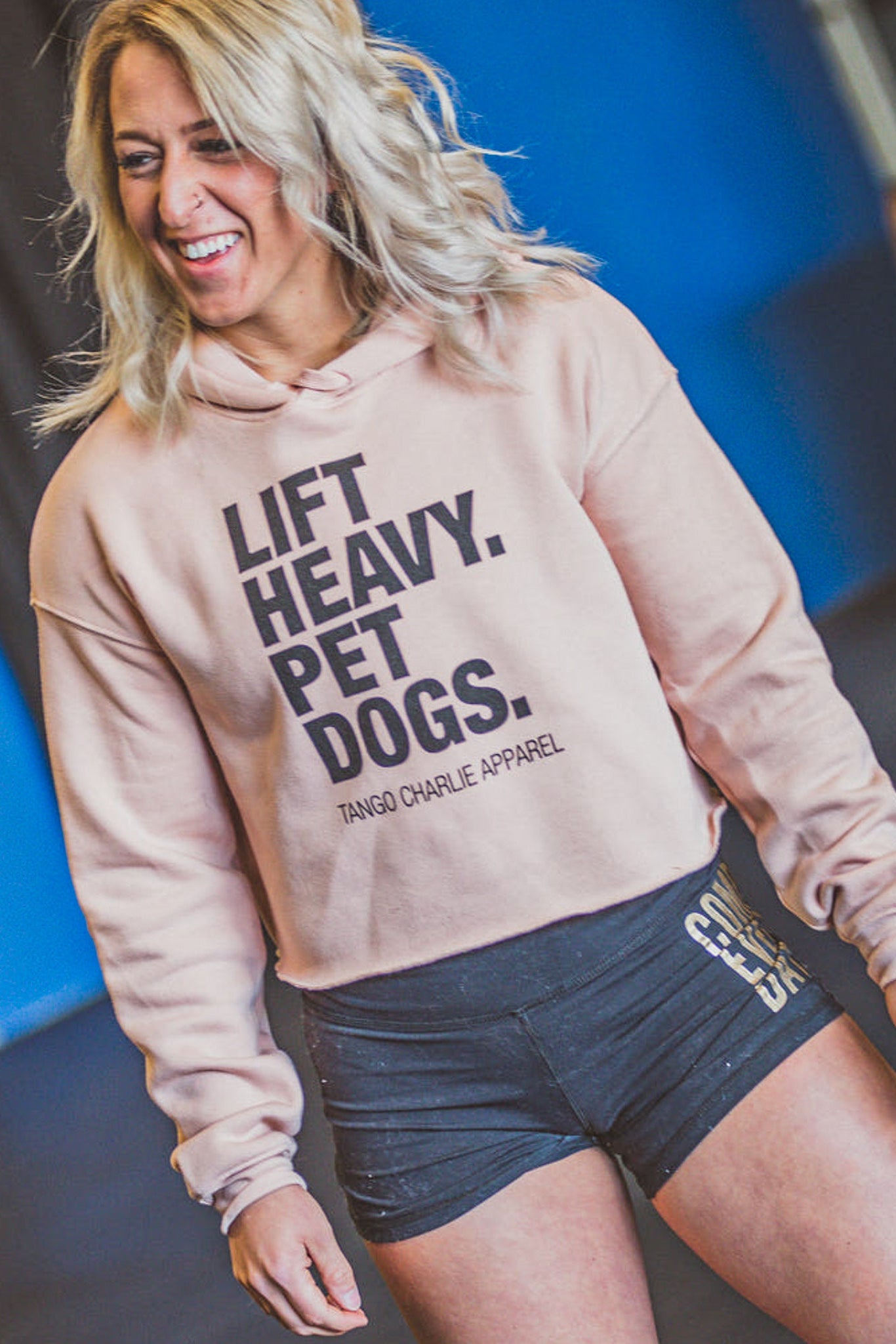 A smiling blonde woman stands indoors, wearing the Tango Charlie Apparel "Lift Heavy. Pet Dogs." Crop Hoodie and black shorts, at what appears to be a gym.