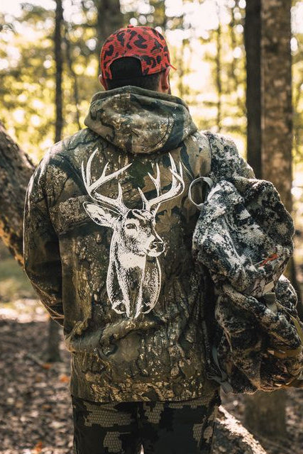 A person wearing The Rut Realtree Premium Hoodie with a large white deer buck graphic on the back, carrying a camouflage backpack through a wooded area.
