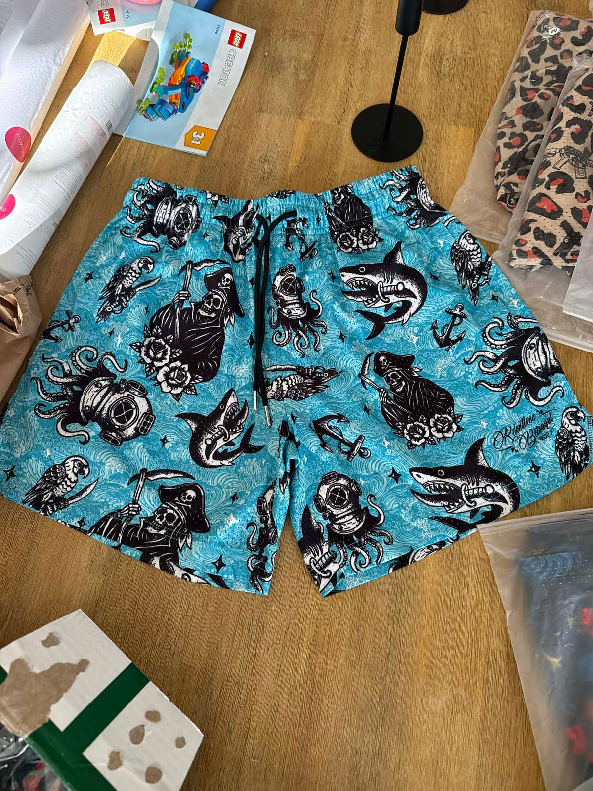 Relentless Betrayal Premium Mesh Shorts | 5.5" - Davy Jones: blue swim trunks featuring black and white tattoo-style mermaids, sharks, anchors, and pirate motifs—displayed flat on a wooden surface.