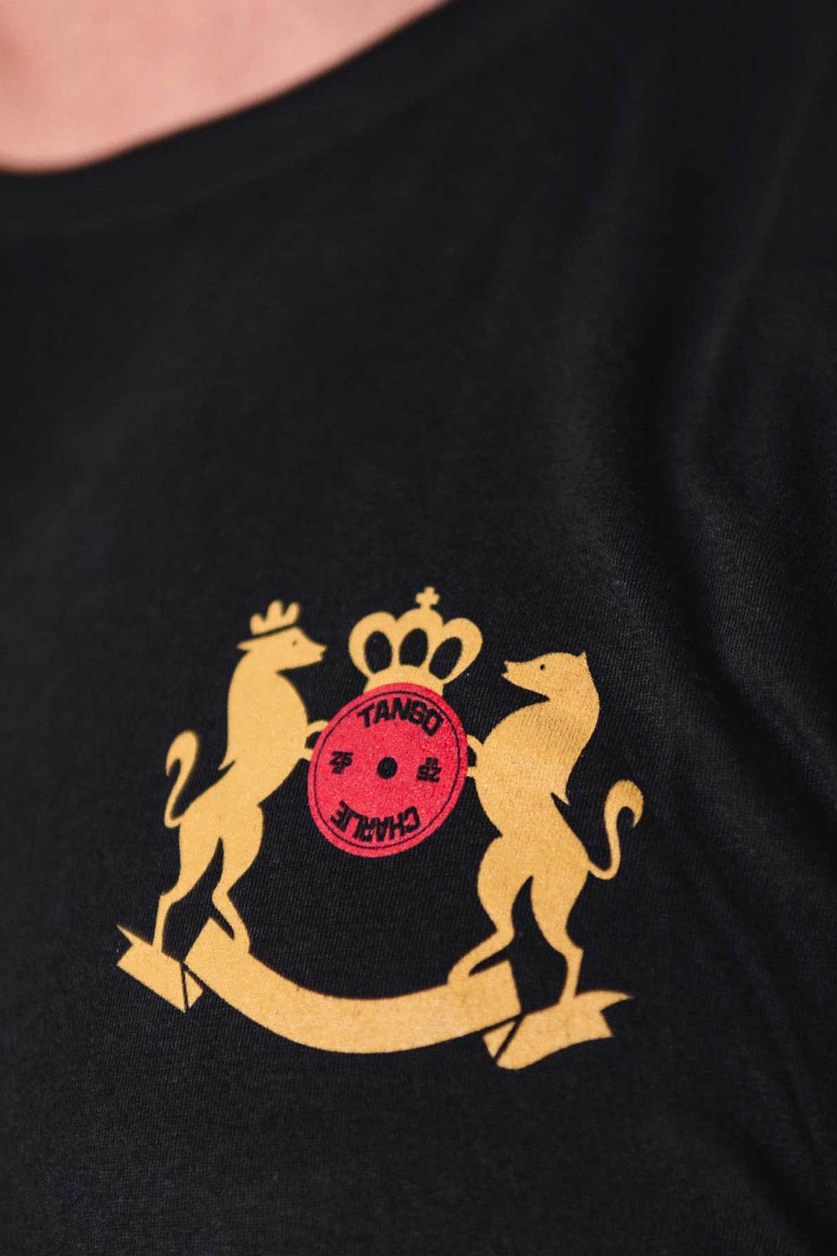 Close-up of the Deadlift Addiction - Tee by Tango Charlie Apparel, a black T-shirt with a gold emblem showing two stylized animals and a crown around a red circle featuring symbols and TANEO JAPAN. Perfect for anyone who takes the lifter generals warning seriously, its as essential as your next deadlift.