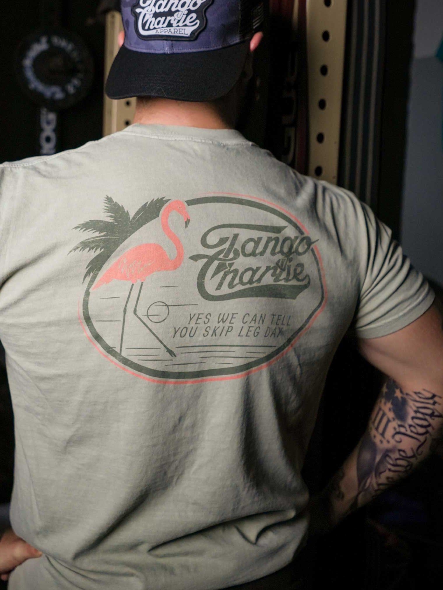 A person in a Tango Charlie Apparel Flamingo Legs Premium Tee and cap stands with their back to the camera. The beige shirt displays a pink flamingo, palm tree, and the text: YES WE CAN TELL YOU SKIP LEG DAY.