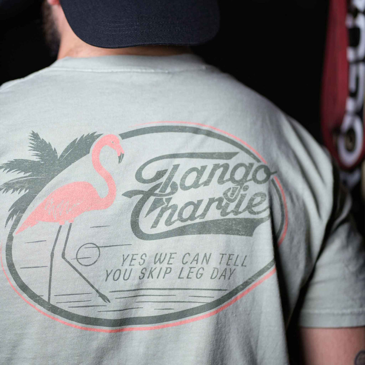 A person wearing the Flamingo Legs - Premium Tee by Tango Charlie Apparel, a breathable light-colored shirt with a flamingo, palm tree, and YES WE CAN TELL YOU SKIP LEG DAY printed on the back.