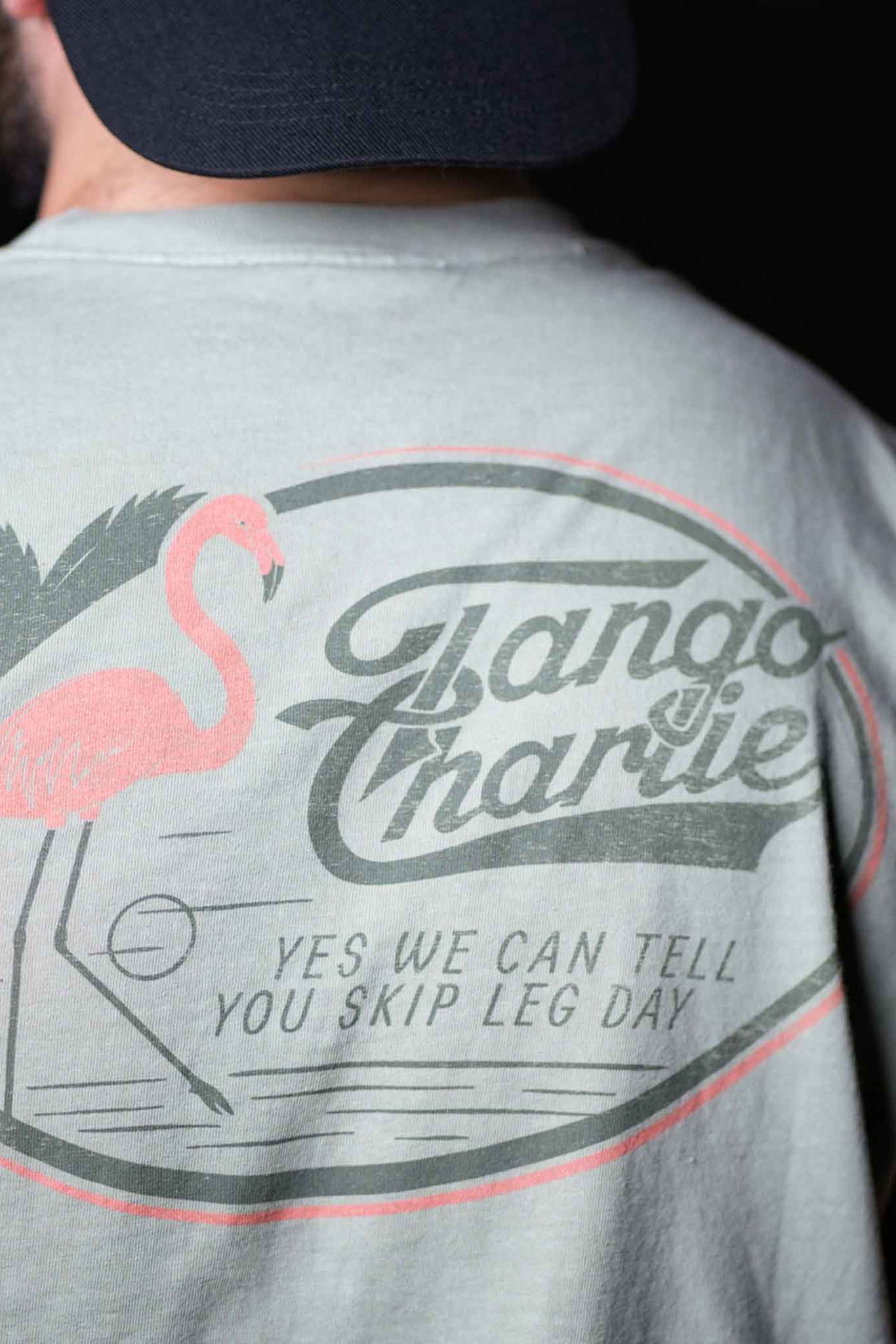 A person wearing the Flamingo Legs - Premium Tee by Tango Charlie Apparel, a breathable light-colored shirt with a flamingo, palm tree, and YES WE CAN TELL YOU SKIP LEG DAY printed on the back.