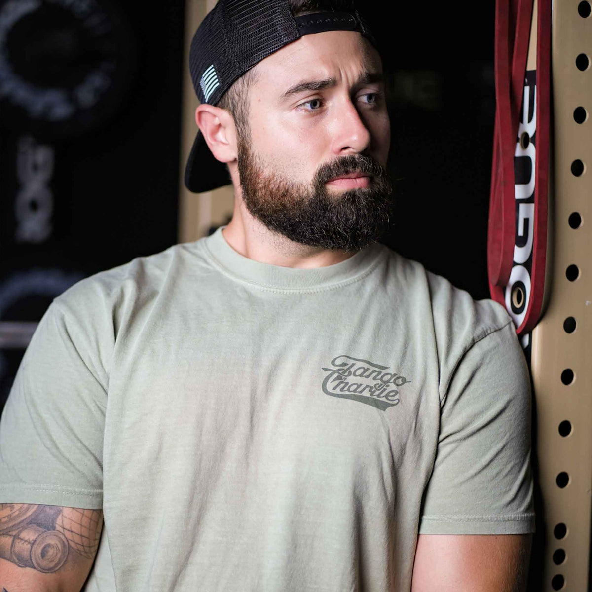 A bearded man in a backward cap leans against a perforated metal wall at the gym, wearing the Flamingo Legs - Premium Tee by Tango Charlie Apparel.