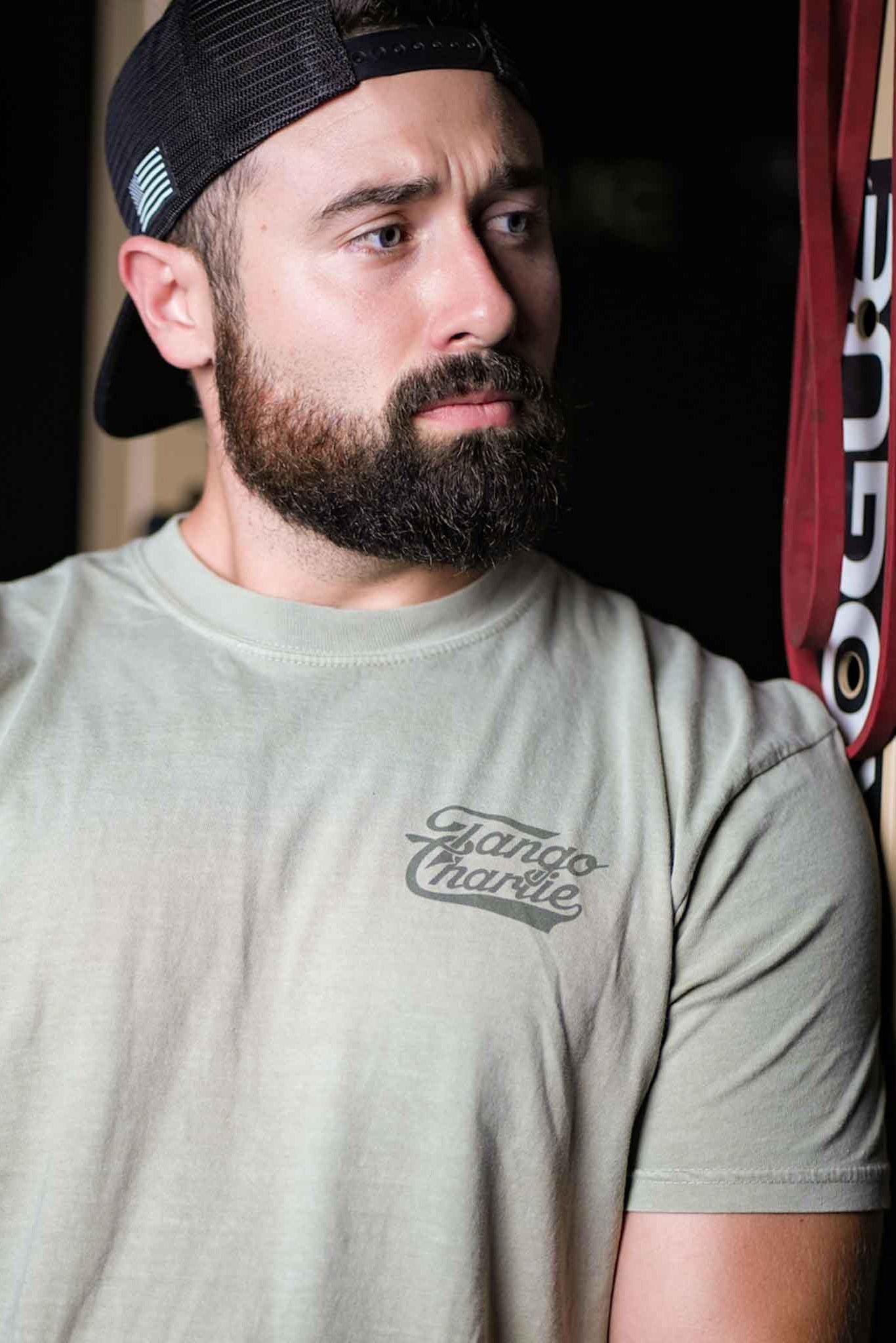 A bearded man in a backward cap leans against a perforated metal wall at the gym, wearing the Flamingo Legs - Premium Tee by Tango Charlie Apparel.