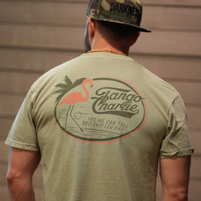 A man faces away from the camera in a Tango Charlie Apparel camo cap and their Flamingo Legs Premium Tee, featuring a flamingo and palm tree with Yes we can tell you skip leg day on the back. This breathable shirt blends comfort and style.