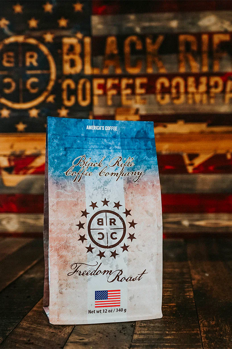 A 12 oz bag of Black Rifle Coffees Freedom Roast Ground sits on a wooden surface with an American flag backdrop, featuring milk chocolate notes, a small US flag, and the Black Rifle Coffee logo.