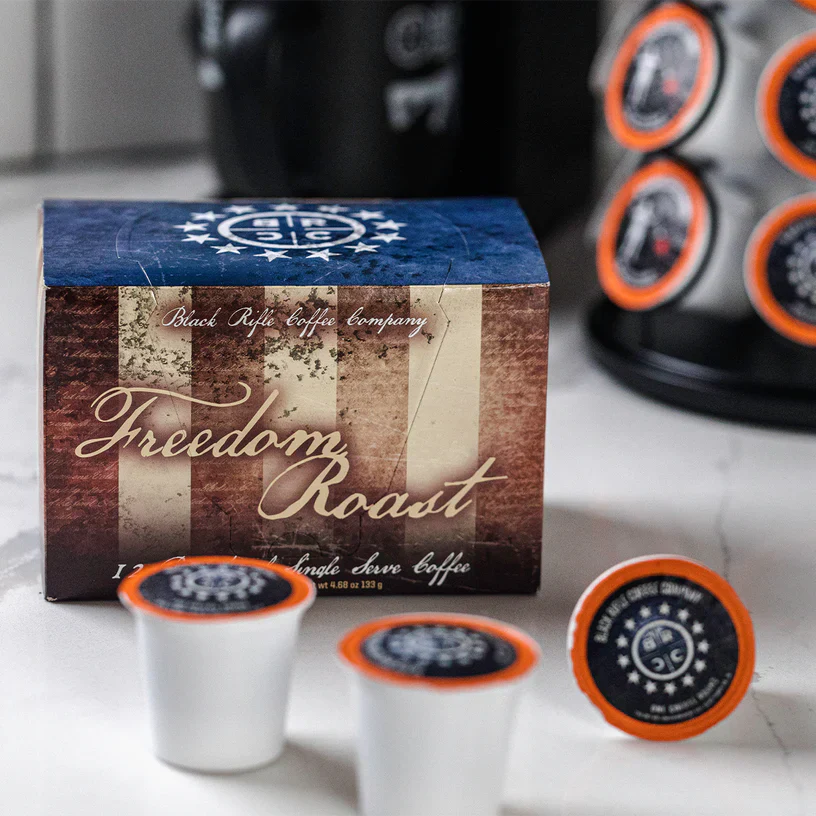 A box of Black Rifle Coffee Freedom Roast Rounds 12 Count sits on the counter with two pods in front and a pod carousel behind it; the box displays an American flag design.