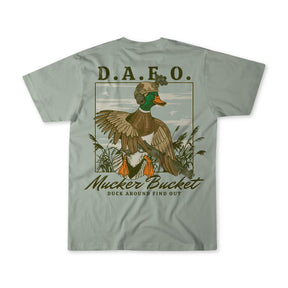 The DAFO Premium Tee from Mucker Bucket is a green T-shirt with a duck in military gear holding a rifle. It features D.A.F.O. above and Mucker Bucket with Duck Around Find Out below, set against reeds and grasses.