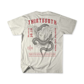 The Serpents And Doves Premium Tee by 13th is a beige T-shirt featuring a winged serpent with a skull. It displays red THIRTEENTH, the date 10.16, and the phrase “Between the Serpents and Doves,” inspired by Matthew 10:16 for wisdom and innocence.