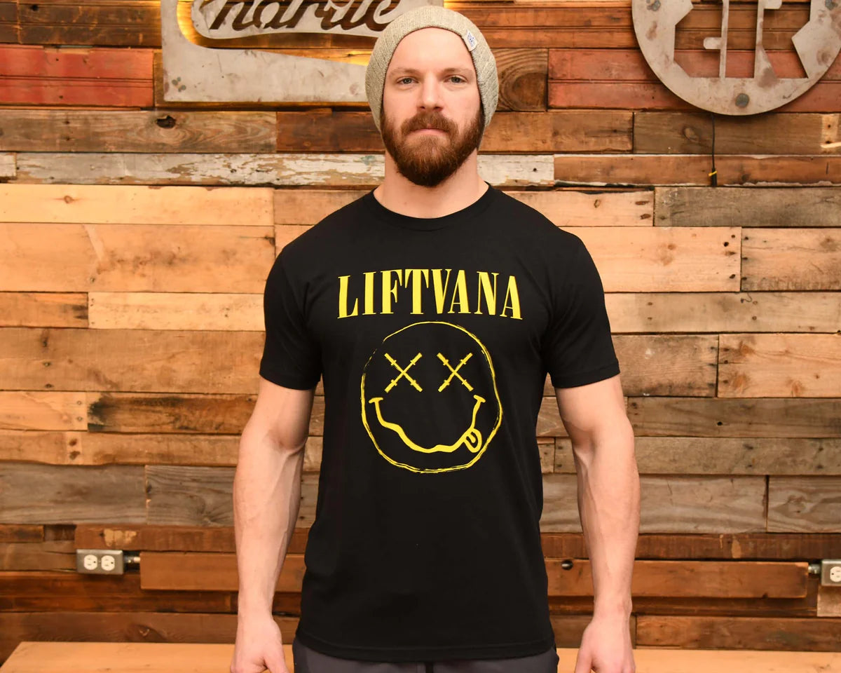 A bearded man in a gray knit beanie and a black "Liftvana" Men's Tee by Tango Charlie Apparel, featuring a yellow smiley face, stands before a wooden plank wall exuding grunge vibes and lifter spirit.