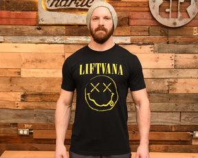 A bearded man in a gray knit beanie and a black "Liftvana" Men's Tee by Tango Charlie Apparel, featuring a yellow smiley face, stands before a wooden plank wall exuding grunge vibes and lifter spirit.