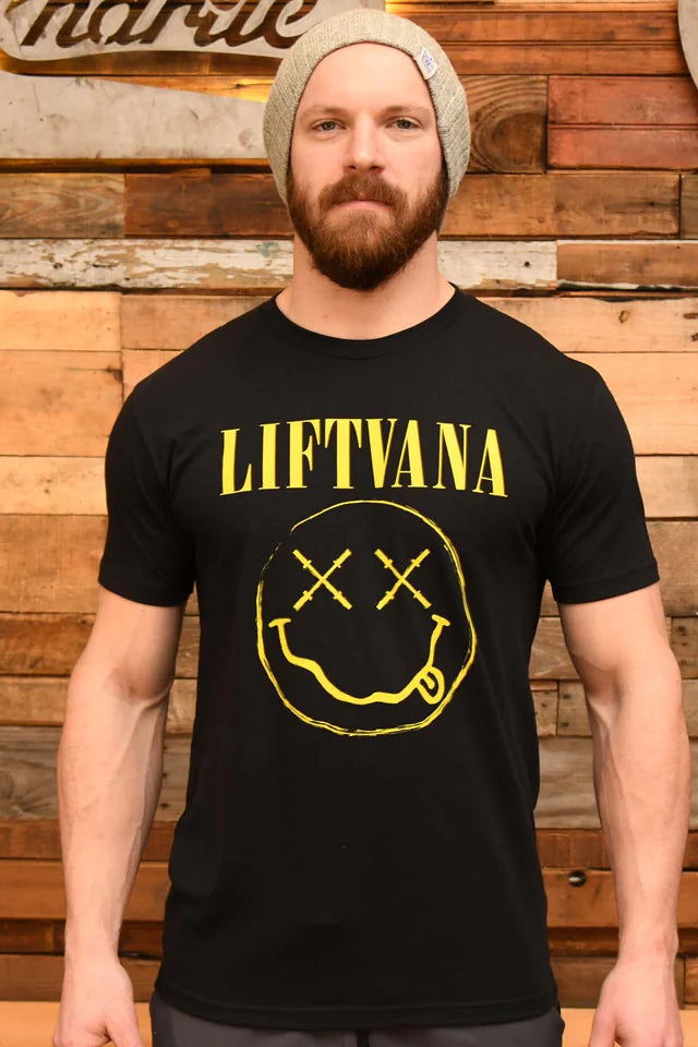 A bearded man in a gray knit beanie and a black "Liftvana" Men's Tee by Tango Charlie Apparel, featuring a yellow smiley face, stands before a wooden plank wall exuding grunge vibes and lifter spirit.