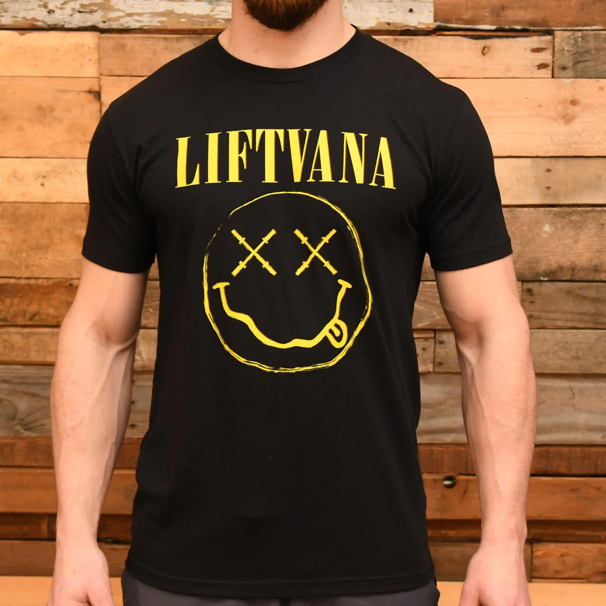 A man wears the "Liftvana" Men's Tee by Tango Charlie Apparel, featuring a smiley face with X eyes and tongue out—a grunge-inspired nod to Nirvana—set against a wooden plank backdrop for an edgy, retro vibe.