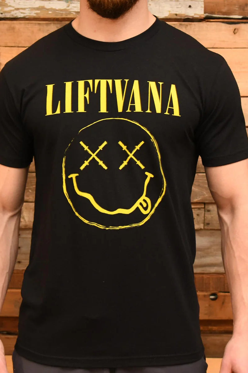 A man wears the "Liftvana" Men's Tee by Tango Charlie Apparel, featuring a smiley face with X eyes and tongue out—a grunge-inspired nod to Nirvana—set against a wooden plank backdrop for an edgy, retro vibe.
