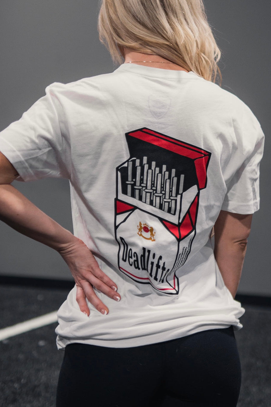 In a gym, a long-haired individual confidently wears the Deadlift Addiction - Tee by Tango Charlie Apparel, featuring the Deadlifts cigarette pack graphic on the back. With a hand on their hip, they embody the lifters warning: deadlifts can be seriously addictive.