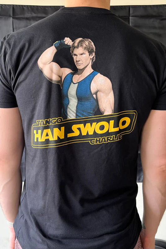 A fit individual dons a Han Swolo T-Shirt from Tango Charlie Apparel, highlighting muscles in a sporty pose. The shirts design resembles retro action movie posters, complemented by the dark backdrop which enhances this dynamic product.