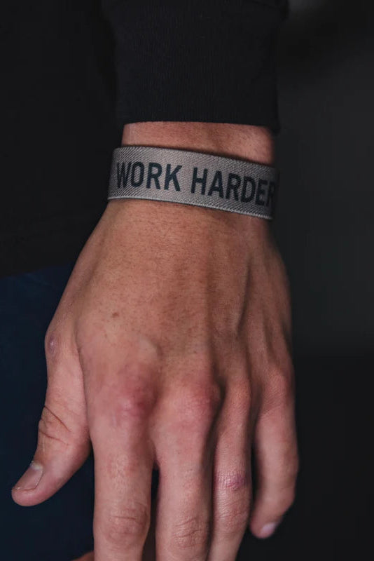 Close-up of a wrist sporting a Tango Charlie Apparels Work Harder Wristband, made from premium fabric. It pairs well with a dark, long-sleeved shirt against a softly blurred background.