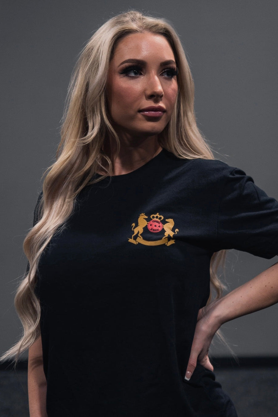 A person with long blonde hair confidently stands with one hand on their hip, dressed in Tango Charlie Apparels Deadlift Addiction Tee, featuring a crest adorned with a crown and lion emblem, against a plain background.