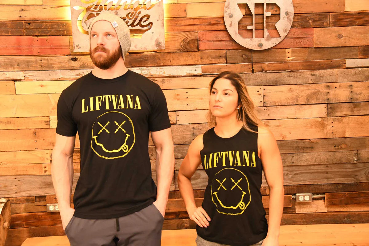 A man and woman stand before a wooden wall, both in black "Liftvana" Men's Tees by Tango Charlie Apparel featuring a yellow graphic. Their serious looks and the man's beanie add grunge energy, reflecting the true lifter’s spirit.