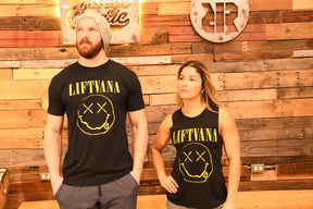 A man and woman stand before a wooden wall, both in black "Liftvana" Men's Tees by Tango Charlie Apparel featuring a yellow graphic. Their serious looks and the man's beanie add grunge energy, reflecting the true lifter’s spirit.