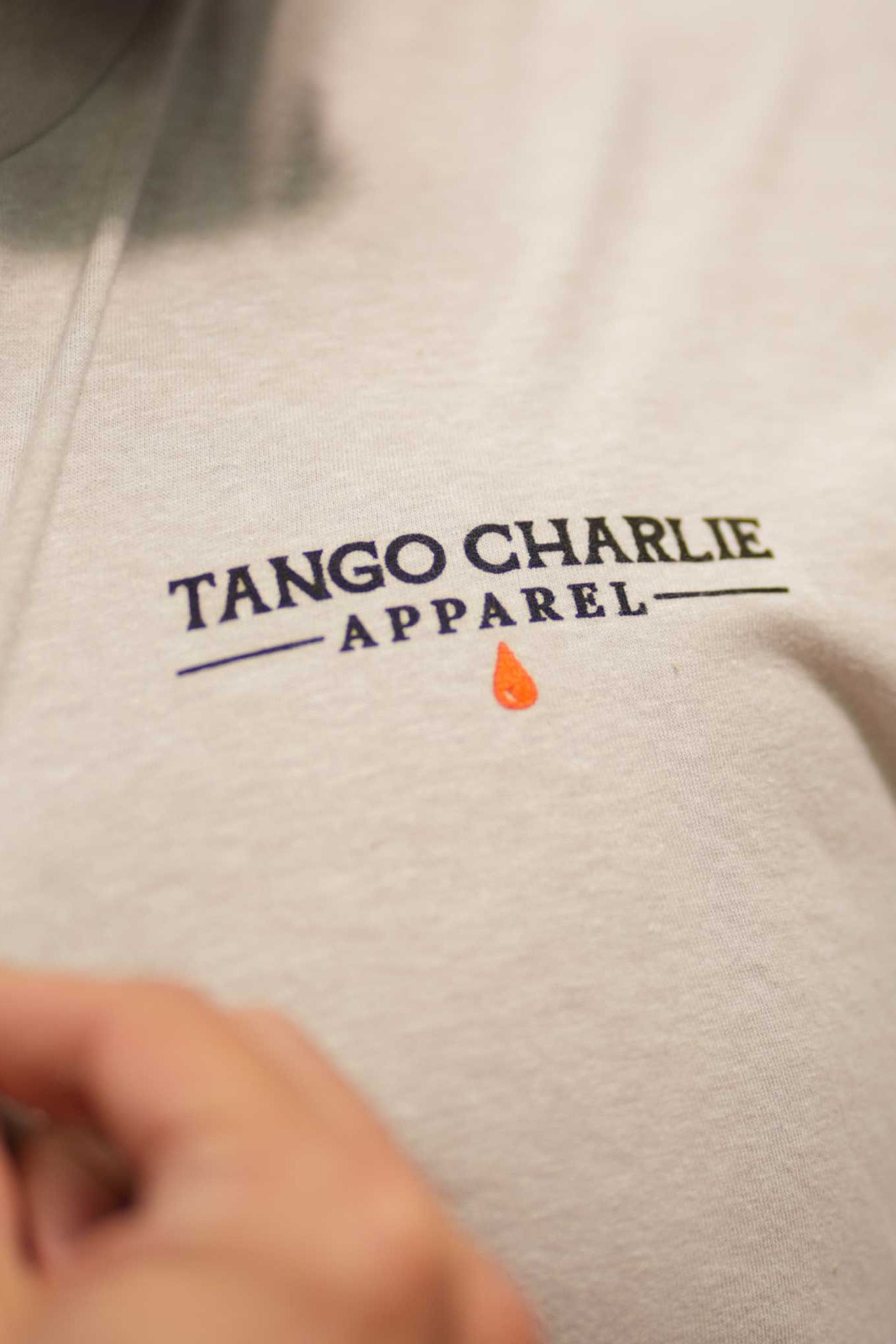 A close-up of someone wearing the Time's Up Tee by Tango Charlie Apparel, made from soft ring-spun cotton. A small orange teardrop sits below the text, as a hand gently holds the shirt near the logo.