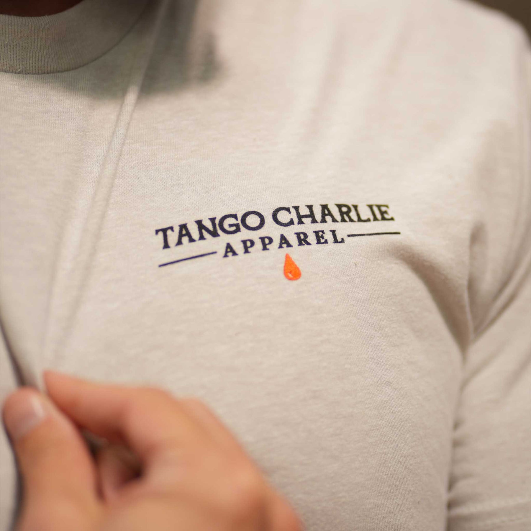 A close-up of someone wearing the Time's Up Tee by Tango Charlie Apparel, made from soft ring-spun cotton. A small orange teardrop sits below the text, as a hand gently holds the shirt near the logo.