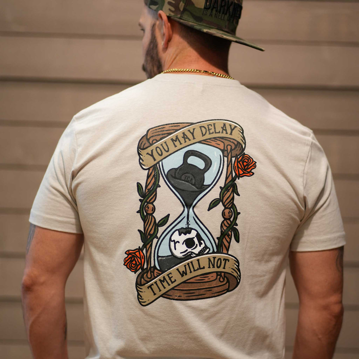 A man wears the Time's Up Tee by Tango Charlie Apparel—a beige workout shirt with an hourglass, kettlebell, and skull graphic on the back. A banner reads, "You may delay, time will not," making it a truly motivational choice.