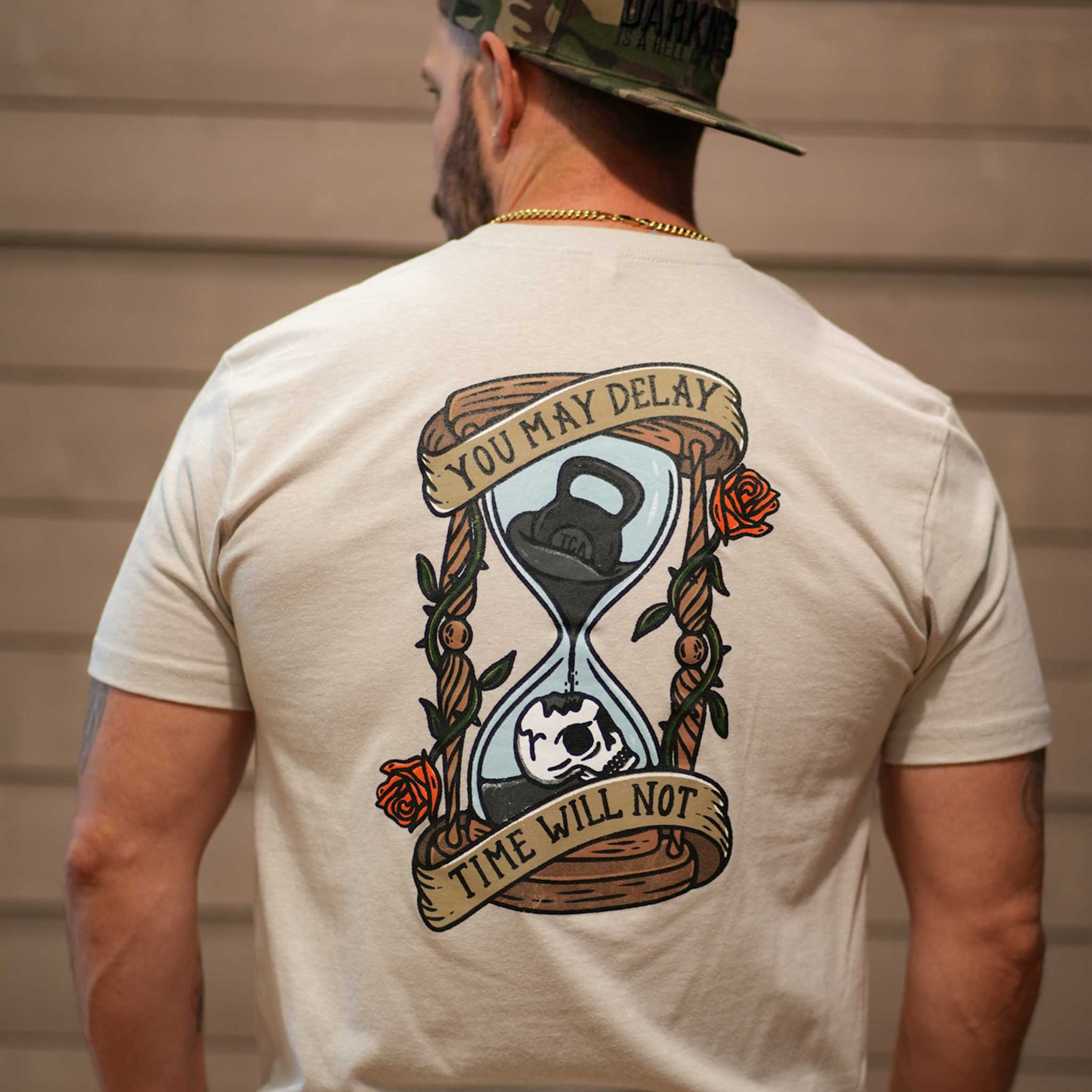 A man wears the Time's Up Tee by Tango Charlie Apparel—a beige workout shirt with an hourglass, kettlebell, and skull graphic on the back. A banner reads, "You may delay, time will not," making it a truly motivational choice.