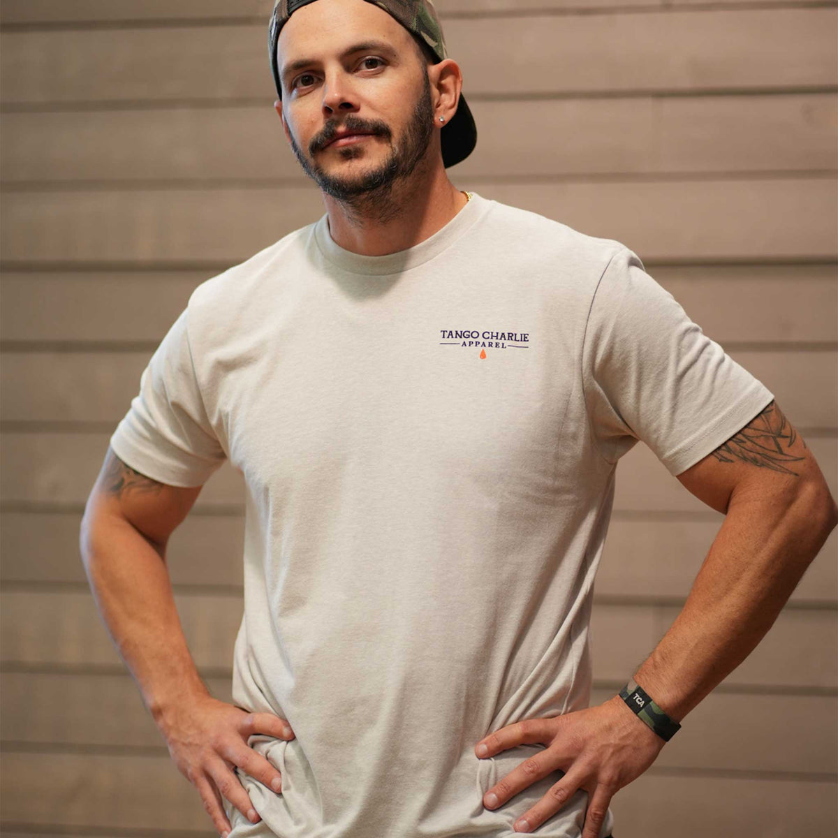 A bearded man in a backward cap stands hands on hips before a wooden wall, wearing the light-colored Time's Up Tee by Tango Charlie Apparel, made from ring-spun cotton and featuring a motivational design.