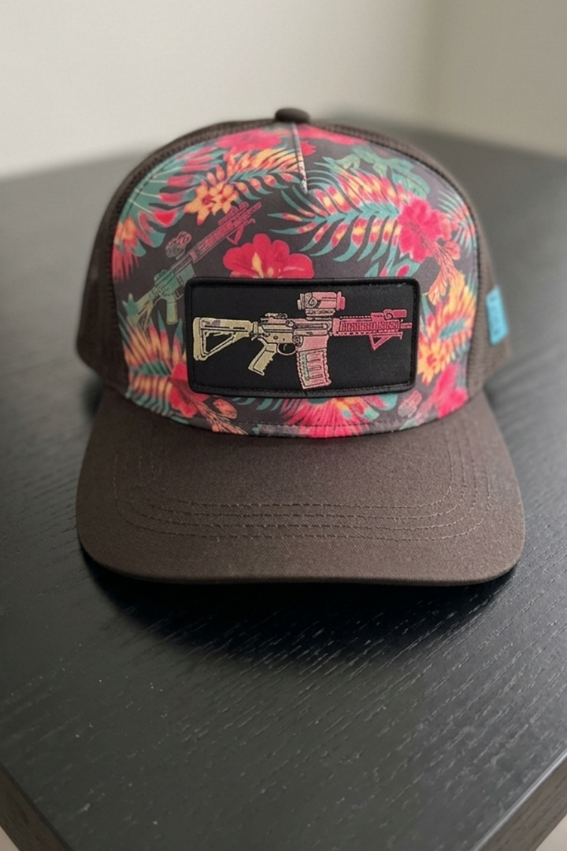 A “Tropical Assassin Hydro” snapback hat by Tango Charlie Apparel, featuring a brown floral design and an illustrated assault rifle patch, rests on the table.