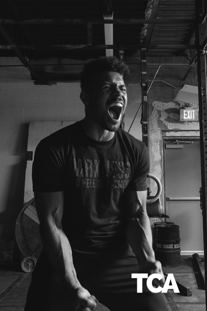 A man in a "Darkness is a HELL of a Coach - Tee" by Tango Charlie Apparel shouts intensely during a gym workout. His muscles are tensed, gym equipment shows in the background, and "TCA" appears at the bottom right of the black-and-white image.