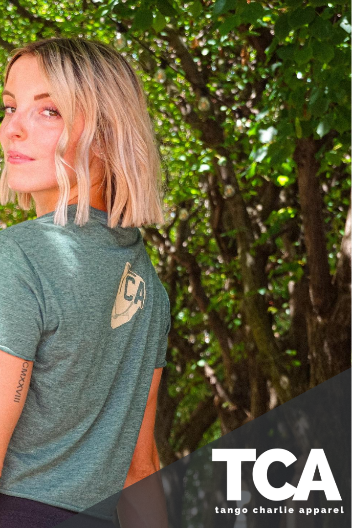 A woman with shoulder-length blonde hair stands outdoors under green trees, wearing a green THICC Boys Fan Club Crop Tee by Tango Charlie Apparel. The TCA (Tango Charlie Apparel) logo appears in the bottom right corner.