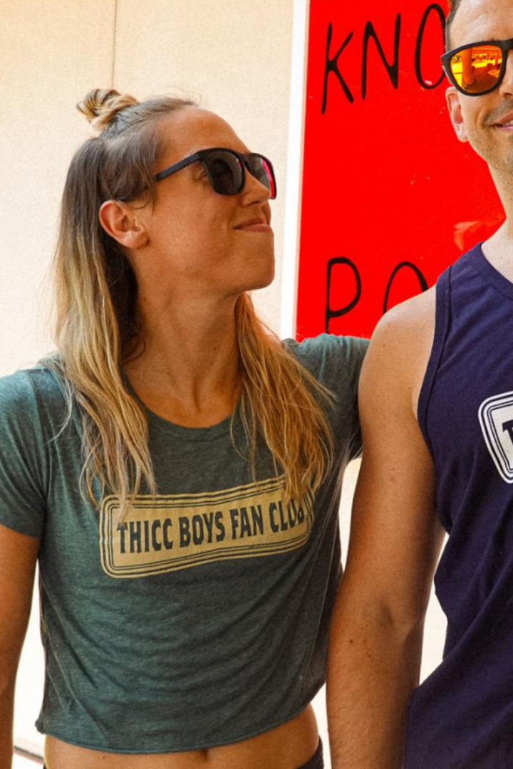 A woman and man in Next Level Brand sunglasses smile together, wearing the Tango Charlie Apparel THICC Boys Fan Club Crop Tee – Green and a THICC Boys Club shirt. The TCA logo appears in the corner; a red sign in back reads “dadbods welcome!”.